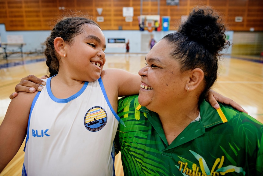 A young girl and her mum smile happily at each other. They are in a basketball court.