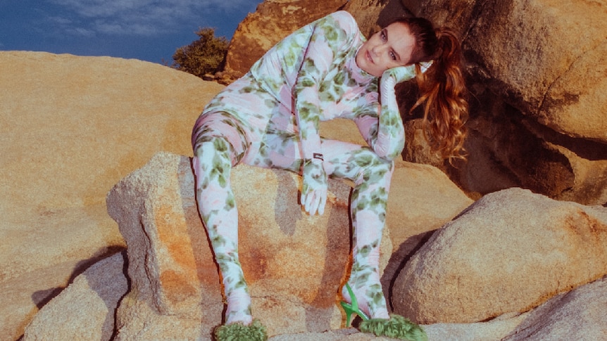 Julia Stone wearing floral bodysuit and green heels, sitting on rocks