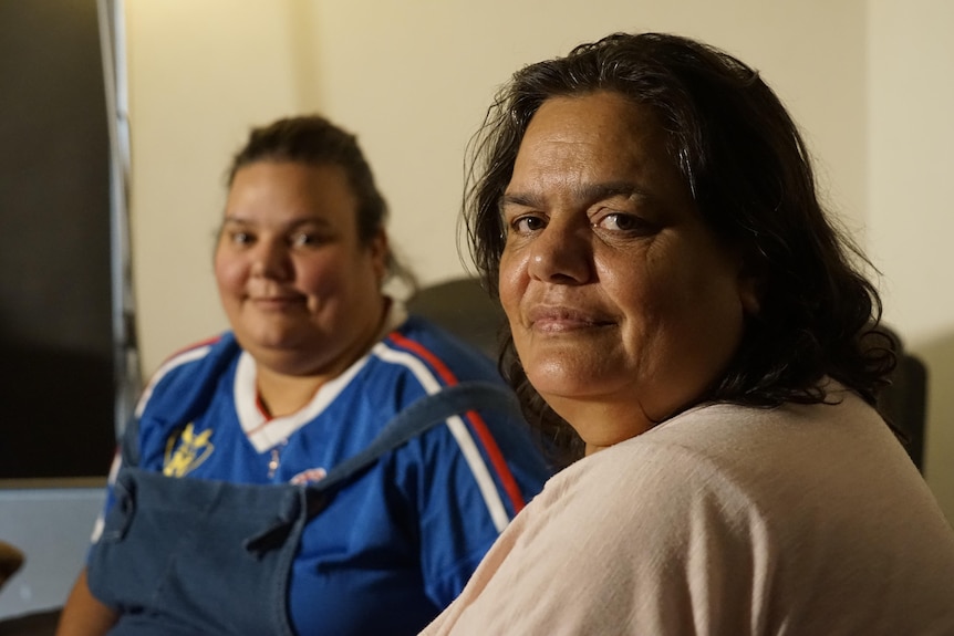 indigenous woman looks over her shoulder at the camera, her daughter sitting behind her