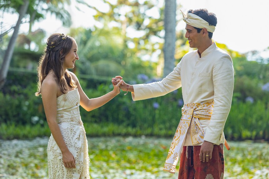 Young white woman with brown hair wears wedding dress and clasps hands with young Indonesian man in white tunic and head scarf.