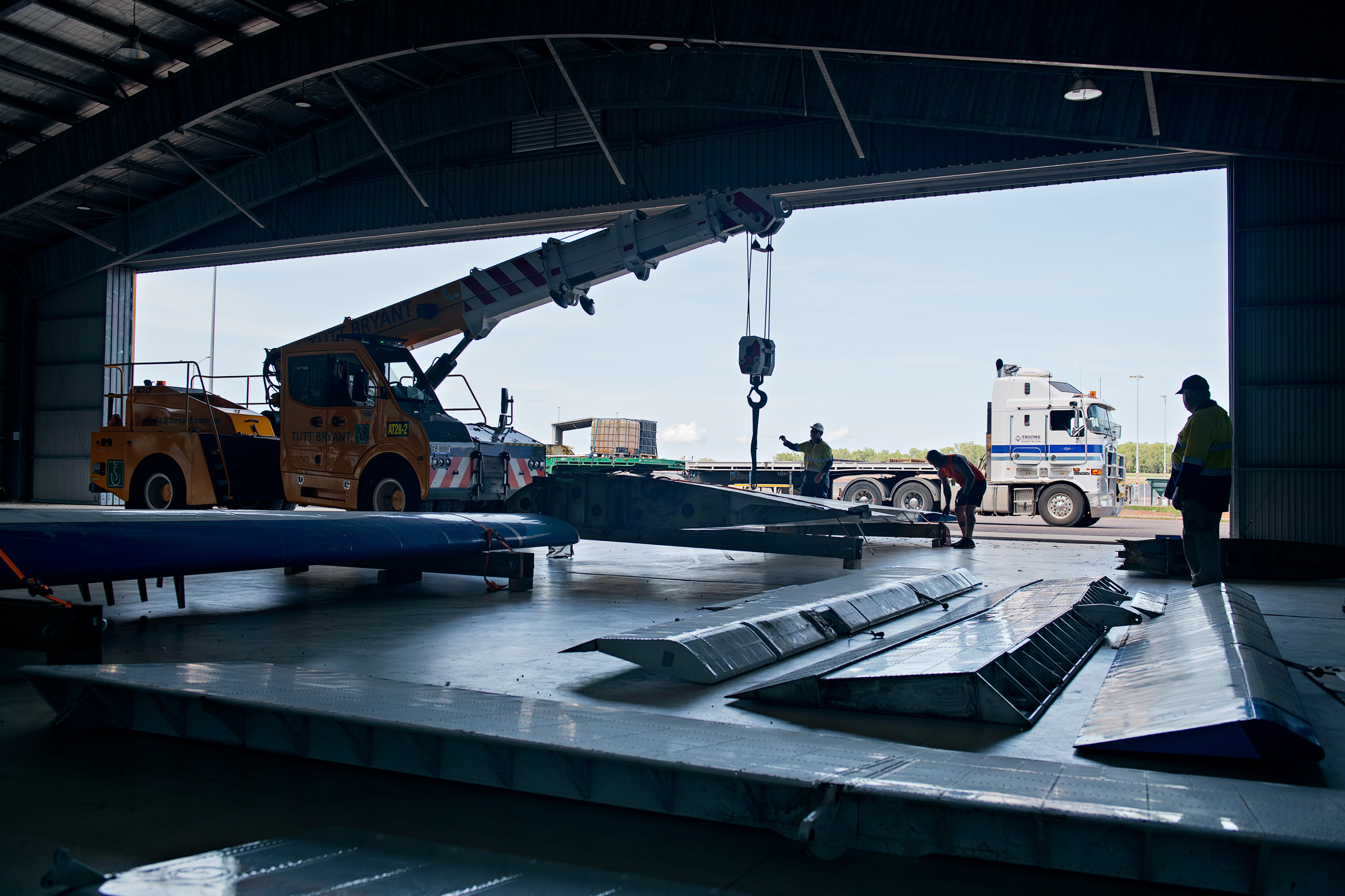 Una imagen sombría tomada en la parte trasera de un cobertizo industrial/hangar de aviones, con piezas de aviones subiendo y trabajadores cerca.