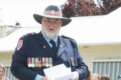 A man with a white beard, wearing uniform, standing near a monument with school children nearby.