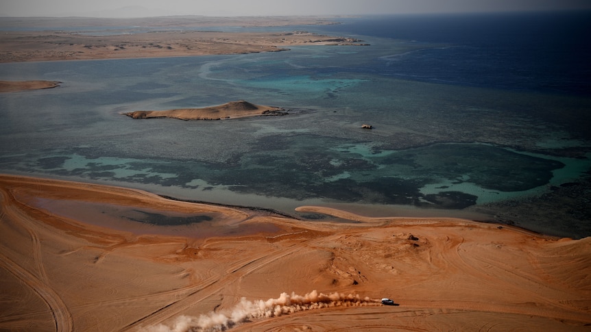 aerial view of red desert with one vehicle making dust clouds, shallow ocean beyond 