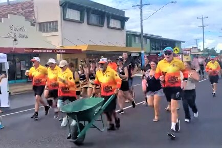 A team of gentleman running together as one pushes a wheelbarrow on the main road in Dimbulah