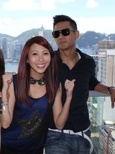 Three people stand on a balcony with a view of Hong Kong, woman in centre pointing to man next to her