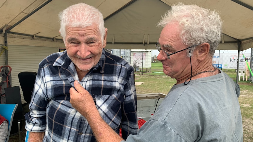 Two grey-haired men, one of whom is smiling, stand underneath an awning.