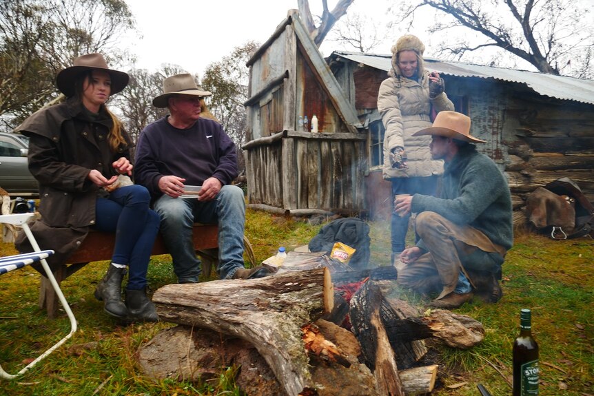 Four people, dressed in wide-brimmed hats and rain jackets, gather around a campfire at a hut in the High Country.