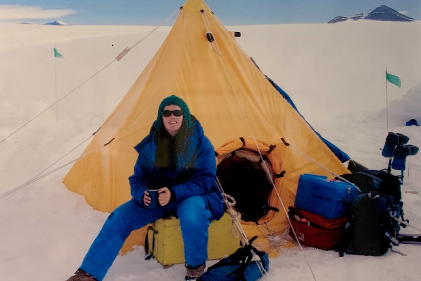 Rachael Mead, in beanie, glasses and thick winter clothing, smiles in front of a yellow tent surrounded by an expanse of ice.