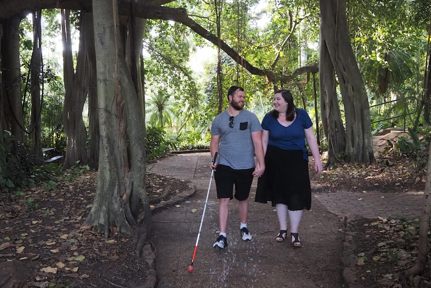 A man using a cane and his fiance holding hands and walking on a path, surrounded by trees. They are smiling at each other.