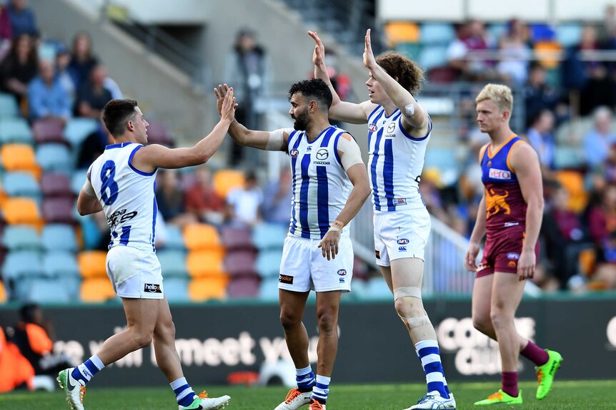Three Kangaroos players high five after kicking a goal as a Brisbane Lions player looks sad in the background.