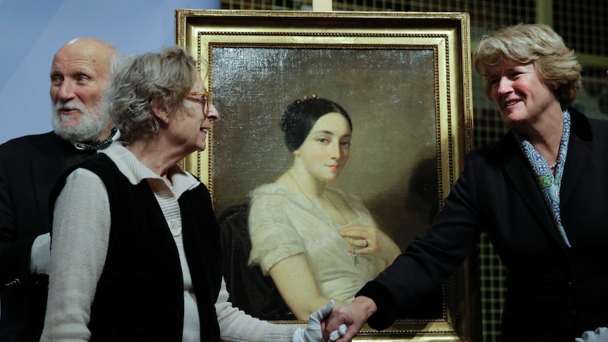 Two women shake hands in front of a painting of a women with black hair.