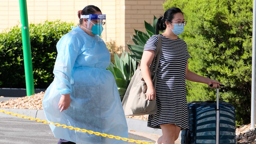 A woman in full PPE escorts a woman with roller luggage.