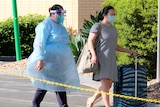 A woman in full PPE escorts a woman with roller luggage.