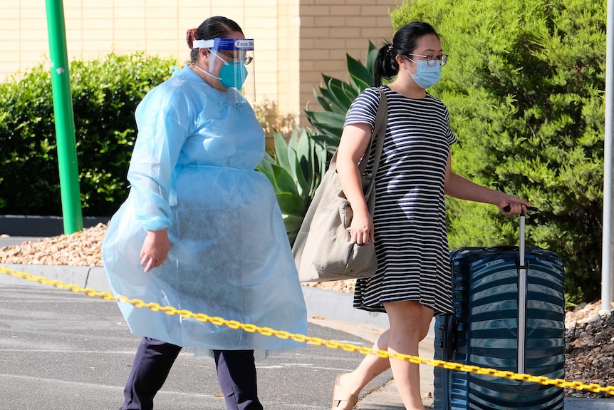 A woman in full PPE escorts a woman with roller luggage.