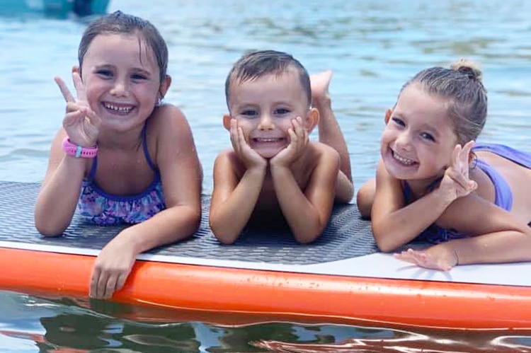 The three children of Hannah and Rowan Baxter, float on a surfboard in the water at a beach.