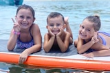 The three children of Hannah and Rowan Baxter, Aaliyah, Trey, and Laianah, float on a surfboard in the water at a beach.