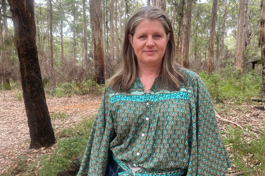 A woman in a green patterned shirt stands among trees in the bush.