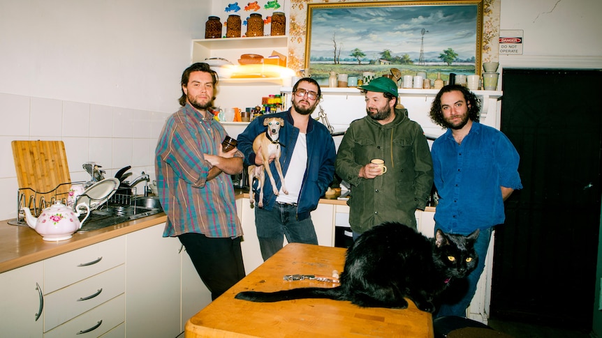 Four men standing side by side in a kitchen. One holds a dog. A black cat is on the table.