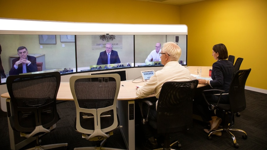 A man and a woman sit at a table and look at three large screens, with men in suits on them.