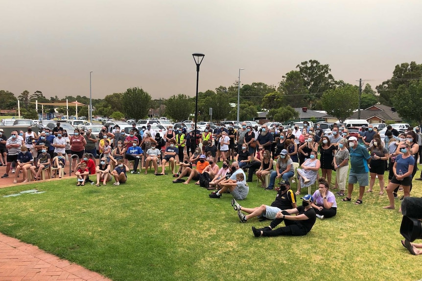 A crowd of residents gather in a park against a smoke filled sky