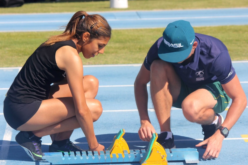 Athlete Ella Pardy and coach Sebastian Kuzminski lean down next to a set of starting blocks on a running track.