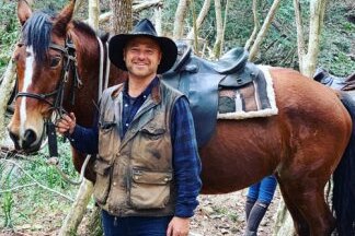 A man in a safari vest and akubra hat holds the reins of a horse in the bush
