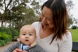 Sue Tay smiles as she cradles her baby son Nathan on her lap on a bench in green parklands.