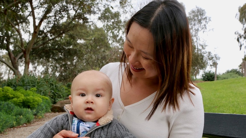 Sue Tay smiles as she cradles her baby son Nathan on her lap on a bench in green parklands.