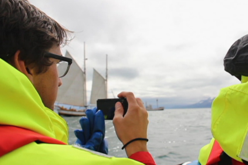 A tight shot from behind two tourists in yellow high-vis jackets looking out to sea, one holding a camera.