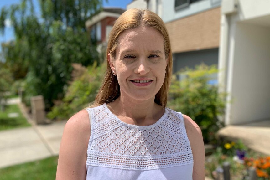 A woman in her 30's with auburn hair wearing a white top, stand outside her house on a sunny day and smiles to the camera.