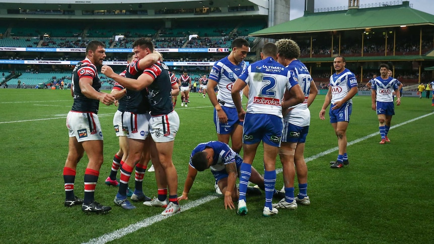 A group of NRL players celebrate in a circle after a try, while their opponents stand around.
