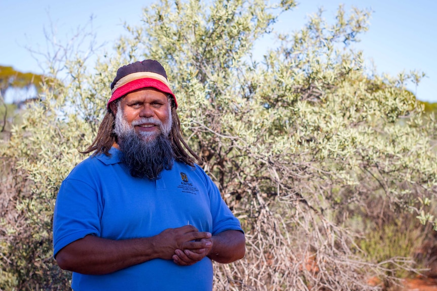 An Aboriginal man with dreadlocks and a beard, wearing a colourful hat, standing in the bush.