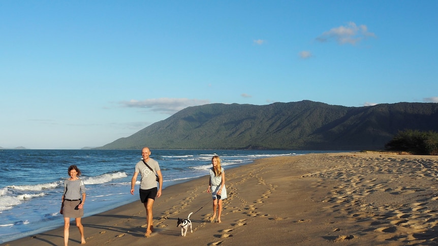 A woman, man and young girl walk along a beach at sunset, with a mountain in the background