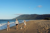 A woman, man and young girl walk along a beach at sunset, with a mountain in the background