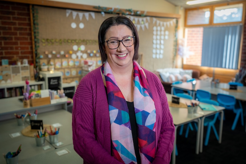 A woman with glasses standards in a classroom, smiling warmly at the camera.