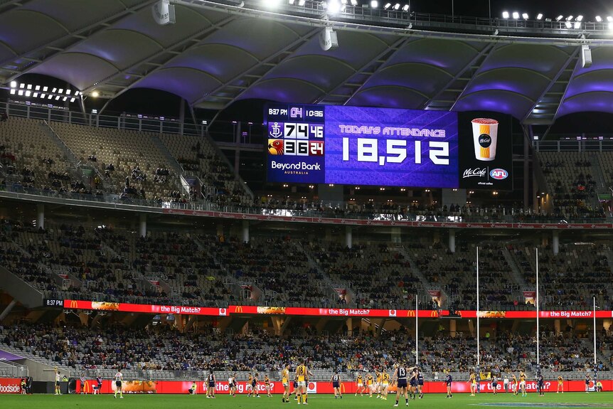 A flood lit stadium shows a number of 18,512 on the screen with AFL players in front of them.