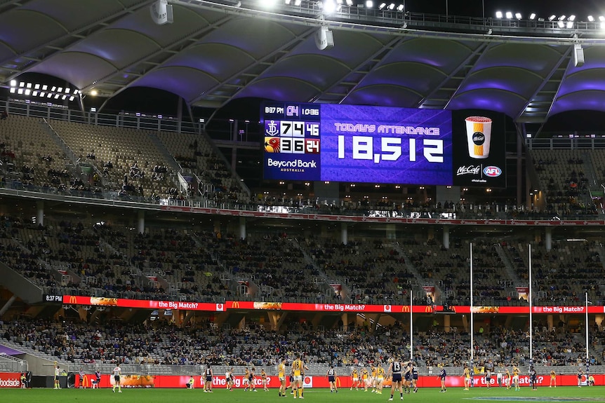 A flood lit stadium shows a number of 18,512 on the screen with AFL players in front of them.