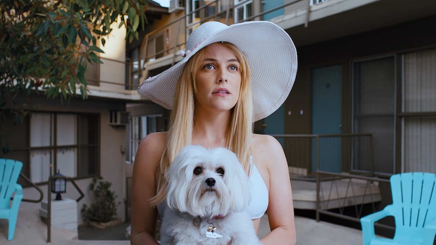 Riley Keogh in a big brimmed hat and carrying a white fluffy dog in the film Under the Silver Lake