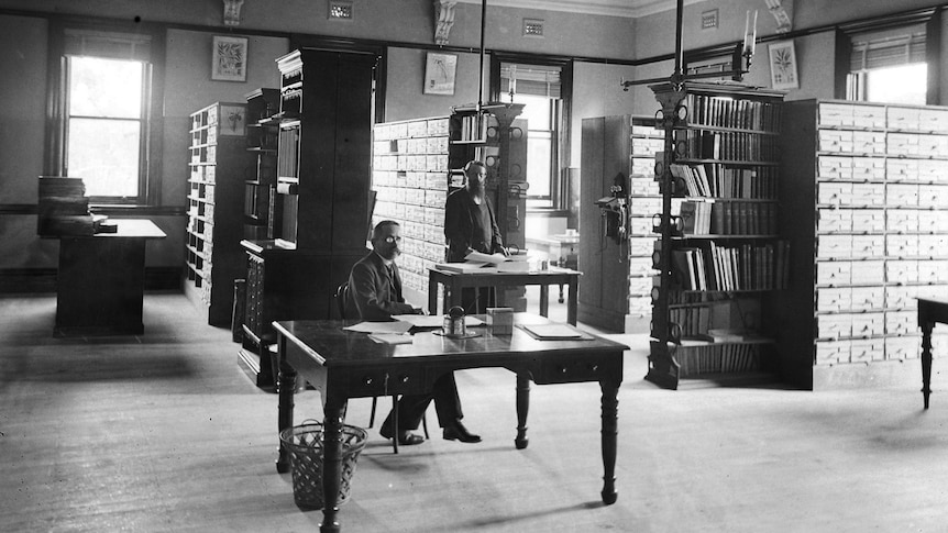 An archival black and white photo of well-dressed men with shelves of boxes