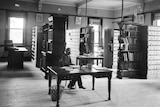 An archival black and white photo of well-dressed men with shelves of boxes