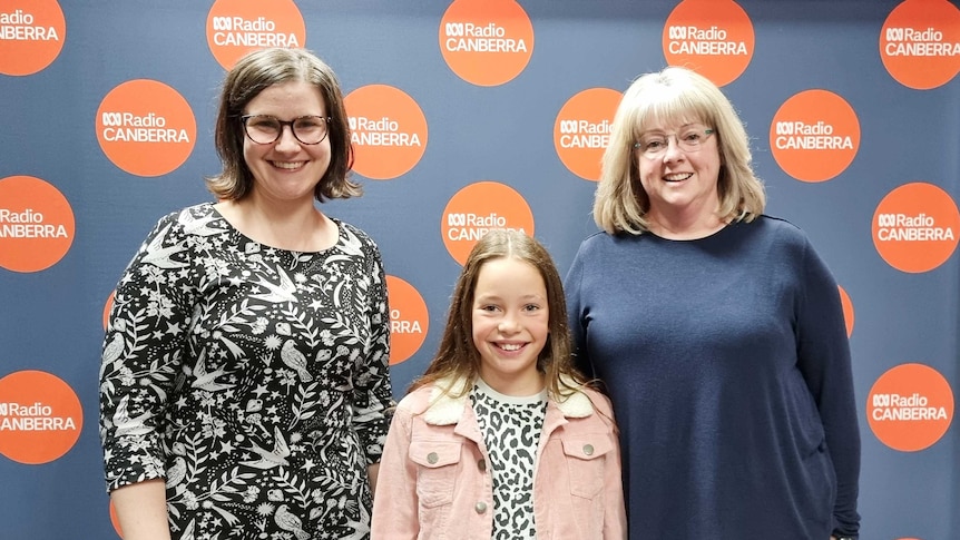 Anna Vidot stands in front of an ABC Canberra banner with Tracy Hall and 9-year-old Lucy Swift in the Northbourne radio studio.