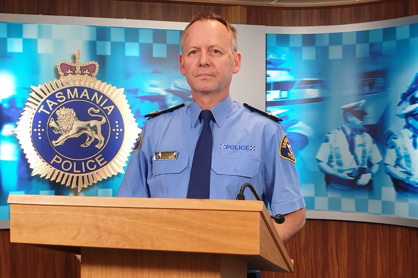 Adrian Bodnar stands at a wooden lectern with large, blue and white Tasmania Police photographs on a wall behind him