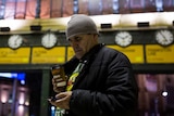 Man stands in front of train station, looking at his phone and holding a coffee cup.