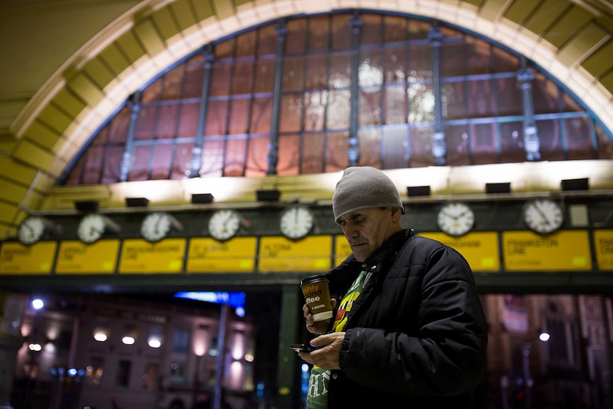 Man stands in front of train station, looking at his phone and holding a coffee cup.