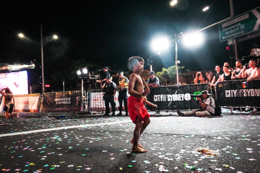 A young Aboriginal boy in red shorts dances with decorated sticks in each hand