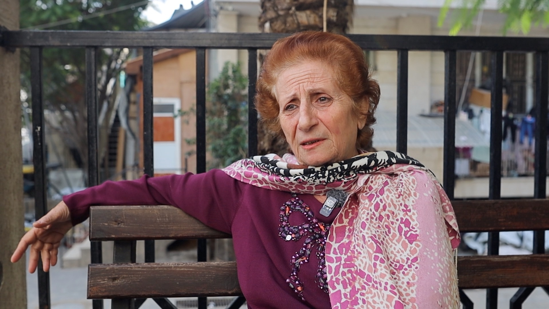 An older red-haired woman with a scarf draped around her neck, sitting on a bench with her arm leaning back 