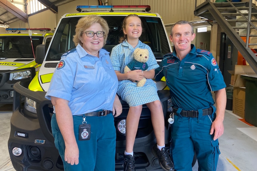Two paramedics standing either side of a young girl who is sitting on an ambulance, holding a teddy bear
