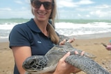 Kim van Oudheusden, holding green sea turtle, smiling, wearing sunglasses, beach, ocean and man in the background.