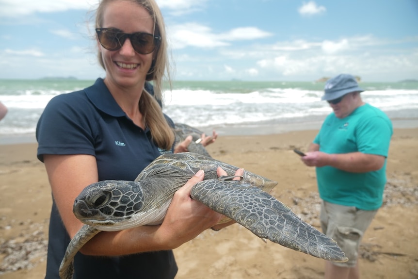 Kim van Oudheusden, holding green sea turtle, smiling, wearing sunglasses, beach, ocean and man in the background.