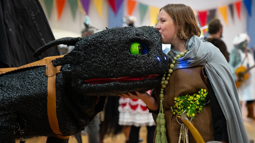 A girl dressed in a grey cape and a brown garment with yellow flowers kisses the head of a black robotic dragon with green eyes.